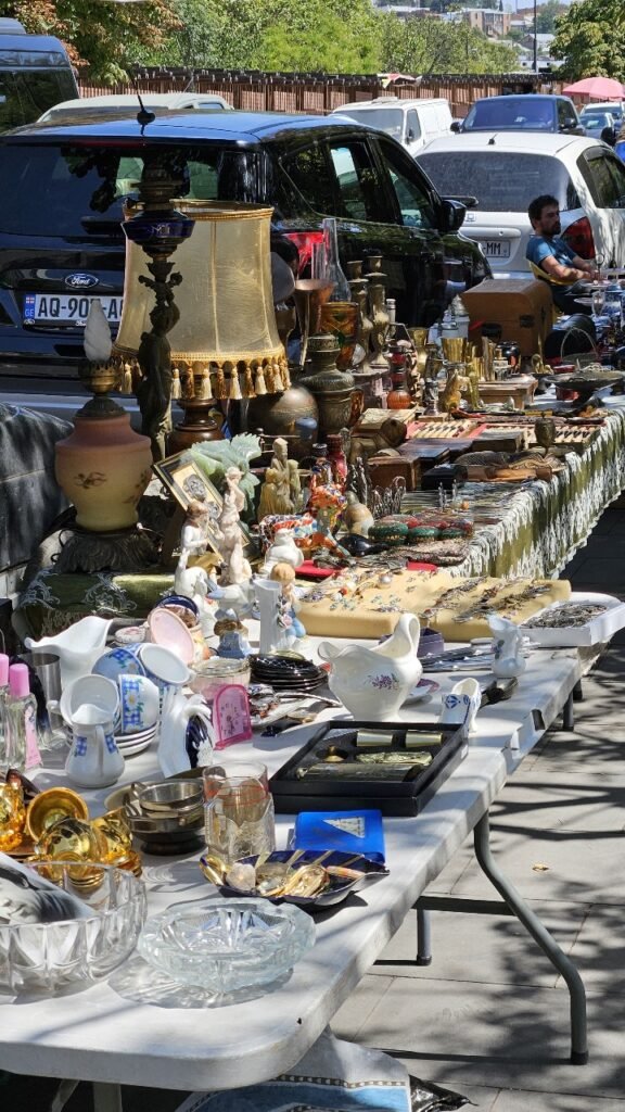 Mesa llena de antigüedades y objetos vintage en el mercadillo al aire libre del Puente Seco de Tbilisi, con coches y árboles en el fondo