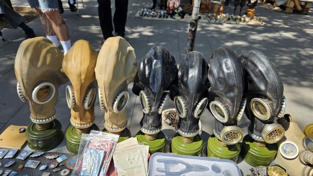 Máscaras de gás vintage em bege e preto alinhadas em um estande do mercado da Ponte Seca de Tbilisi, com emblemas e outros objetos da era soviética em primeiro plano