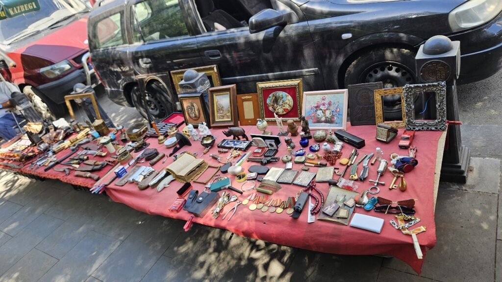 Mesa con mantel rojo repleta de diversos objetos antiguos y curiosidades en el mercadillo del Puente Seco de Tbilisi, con coches estacionados en el fondo