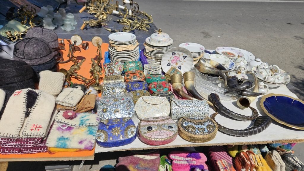 Variedad de antigüedades y artesanías en un puesto del mercadillo del Puente Seco de Tbilisi, incluyendo vajillas, cajas decorativas, sombreros tradicionales y objetos de metal